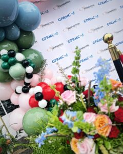 A backdrop with GPRMC logos repeating. There's a balloon tower and flowers in the foreground.